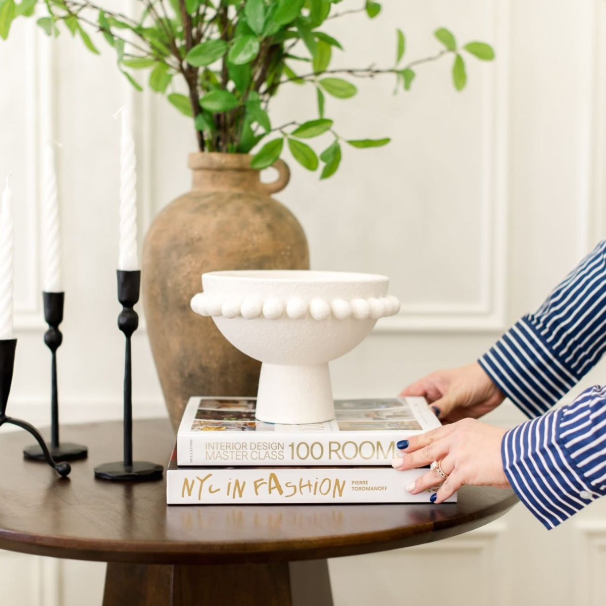Woman's hands stacking books on a table with candle sticks and a vase with greenery behind it